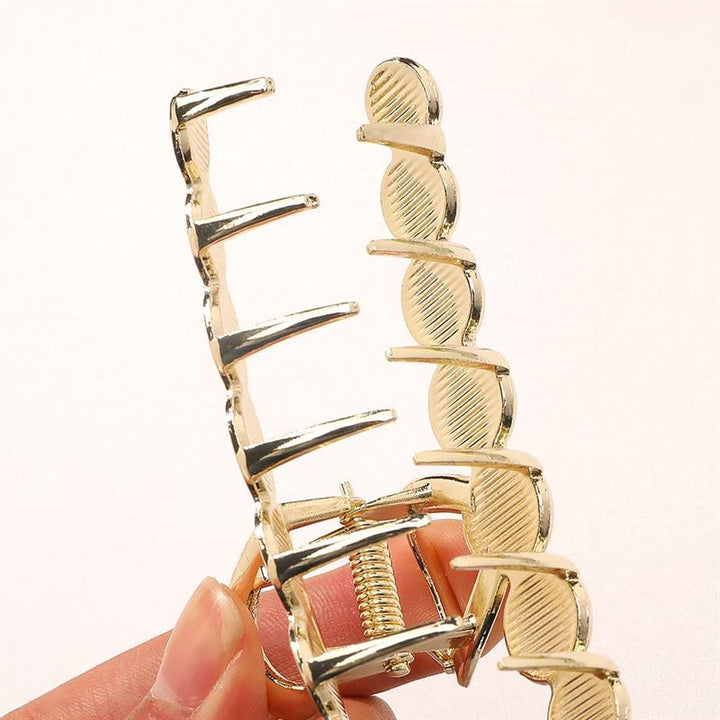 A close-up of a hand holding the SOHO Becca Hair Claw - Midnight by SOHO, featuring a wavy gold-colored metal design and interlocking teeth for a secure messy bun, shown against a plain light background.