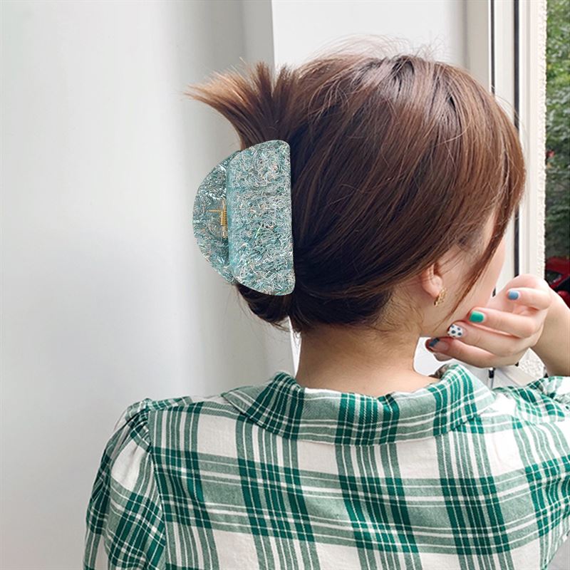 A person with short brown hair, clipped up with the SOHO Cleo Hair Claw in Crystal Blue, gazes outside through a window. They wear a green and white plaid shirt and rest their painted nails against their face.