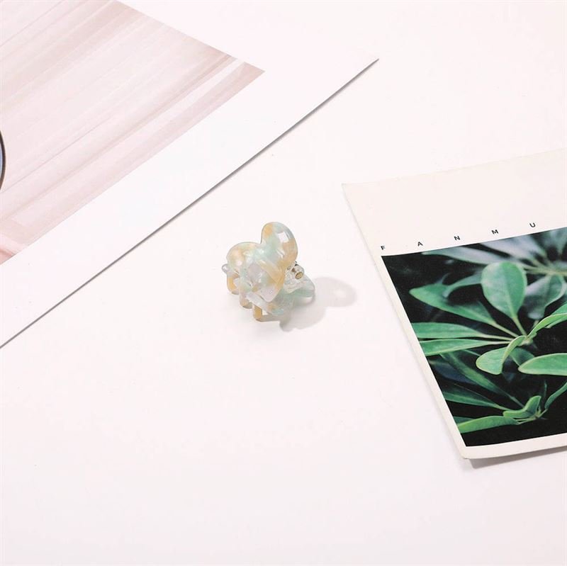 The SOHO Hara Mini Hair Claw - Beach rests on a white surface next to two papers—one with a green leafy plant photo and another with a pale pink abstract pattern—offering a secure everyday hair grip.