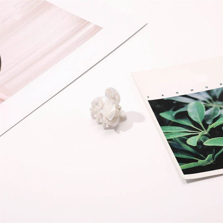 The SOHO Hara Mini Hair Claw - Porcelain by SOHO, a small white translucent clip, rests on a white surface between plant and abstract paper accents, offering a secure hair grip and stylish touch.