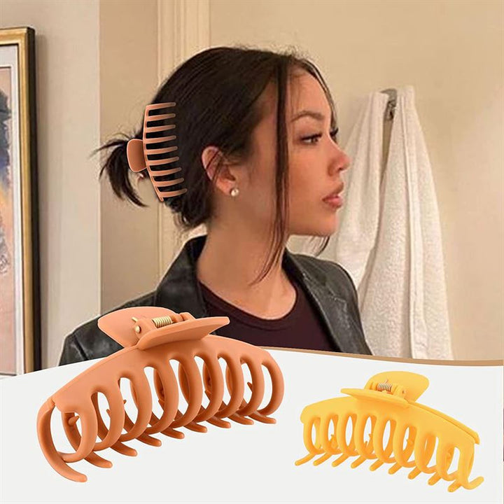 A woman with dark hair in a bun clipped with the SOHO Kana Hair Claw - Bark Brown wears a black jacket. In front are two large SOHO Hair Claws, brown and yellow, for all hair types. A white towel hangs in the background.