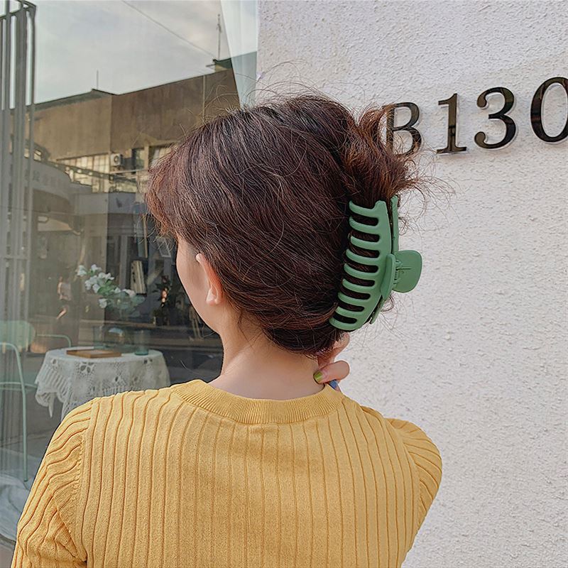 A person with wavy brown hair wears a yellow ribbed top and secures their bun with the SOHO Kana Hair Claw - Bark Brown, suitable for all hair types, while standing outside near a white wall marked B130.