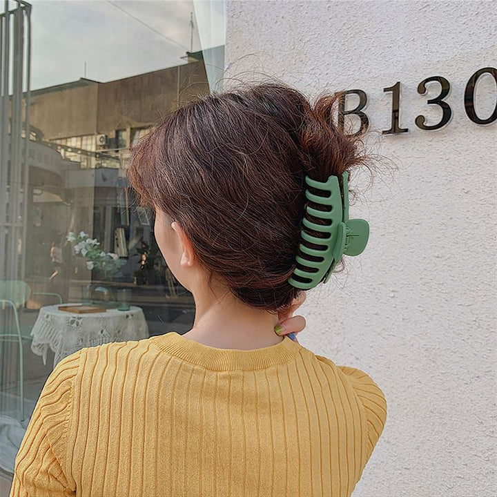 A person with wavy brown hair wears a yellow ribbed top and secures their bun with the SOHO Kana Hair Claw - Bark Brown, suitable for all hair types, while standing outside near a white wall marked B130.