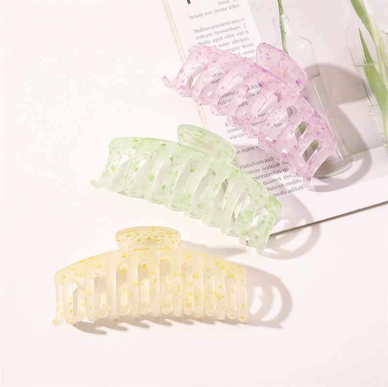 The SOHO Kana Splash Hair Claw in Purple by SOHO is displayed on an open magazine with a glass bottle and plant stem, offering a strong grip and suitable for all hair types against a white background.