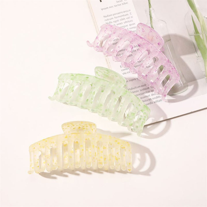 The SOHO Kana Splash Hair Claw in Purple by SOHO is displayed on an open magazine with a glass bottle and plant stem, offering a strong grip and suitable for all hair types against a white background.