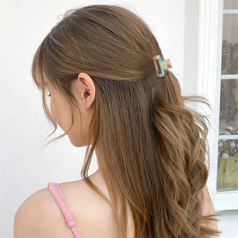 A woman with long, wavy brown hair wears a pink tank top and holds her half-up style with the SOHO Leia Mini Hair Claw - Beige Marble. She faces away from the camera, standing indoors near a window.
