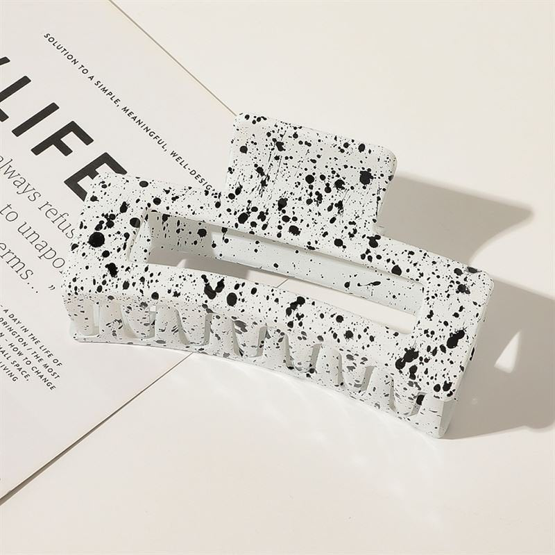 The SOHO Luva Hair Claw - White by SOHO, featuring a bold black splash pattern, rests on a white surface and partially overlaps the corner of a sheet of paper with printed text.