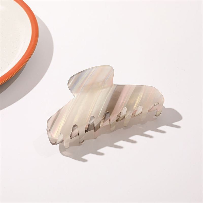 The SOHO Mesa Hair Claw - Grey Rainbow by SOHO, featuring a marbled pearlescent design, sits on a white surface near the edge of a white plate with an orange rim.