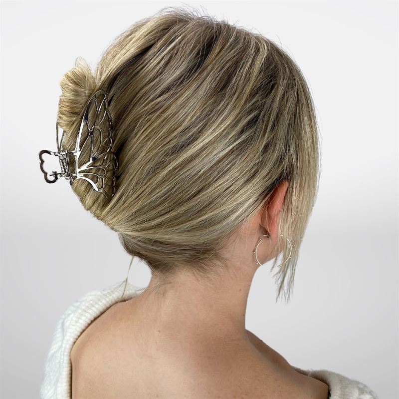 A woman with blonde hair in a neat bun, secured by the SOHO Metal Butterfly Hair Claw - Silver, wears a white top and hoop earrings, shown from the back against a plain background.