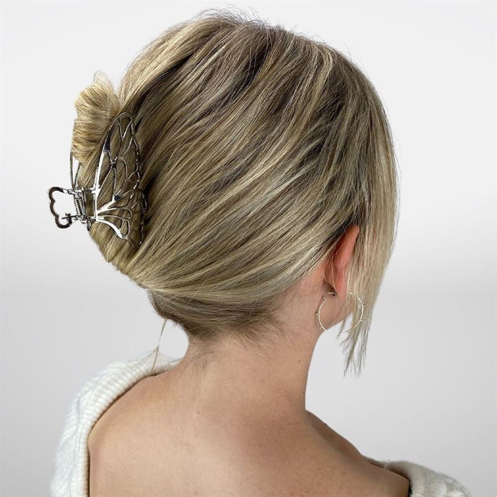 A woman with blonde hair in a neat bun, secured by the SOHO Metal Butterfly Hair Claw - Silver, wears a white top and hoop earrings, shown from the back against a plain background.