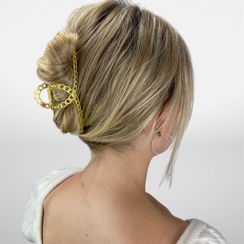 A woman with blonde hair in a low twist, secured by the SOHO Monz Metal Hair Claw in Gold from SOHO. She wears a white sweater and large hoop earrings, with her back to the camera.