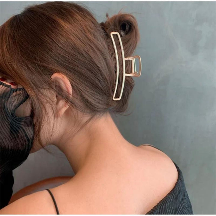 A woman with brown hair styled in a secure messy bun with the SOHO Selin Metal Hair Claw in Silver is seen from behind. She wears a black textured sleeveless top, holding sheer black fabric, suitable for all hair types against a plain gray wall.