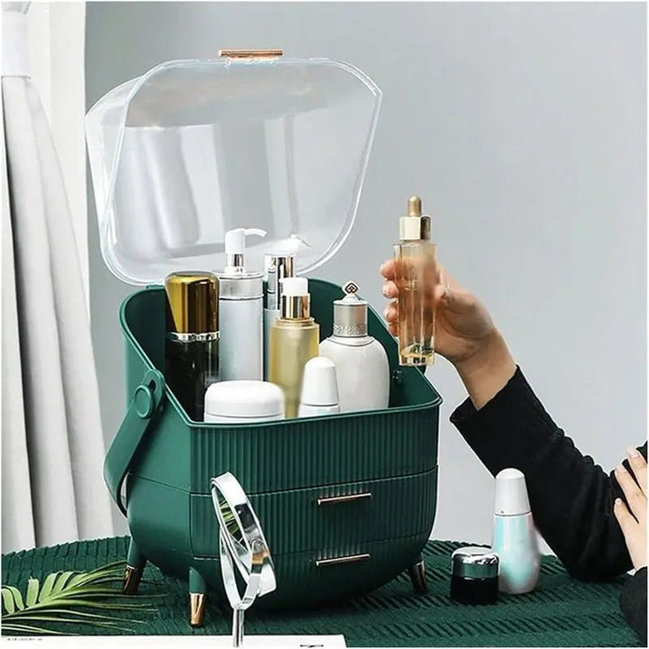 A hand holds a dropper bottle above the UNIQ Retro Makeup Box Organiser by UNIQ in Dark Mint/Dark Green, filled with skincare and beauty items. The organiser has a clear lid and two drawers for neat storage, set on a green table.